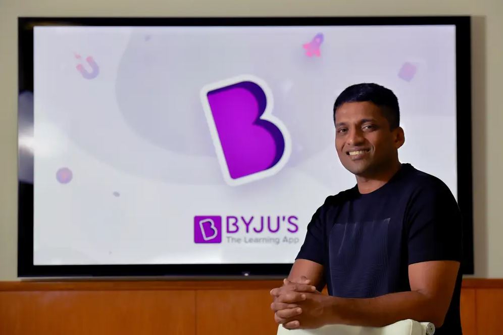 Byju Raveendran sitting in front of a Byju’s logo display amid the company’s rise and fall.