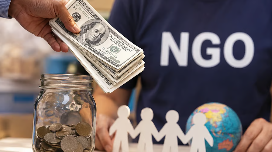 Hand placing US dollar notes into a donation jar beside an NGO volunteer, representing India's 2026 FCRA foreign funding regulations for NGOs.